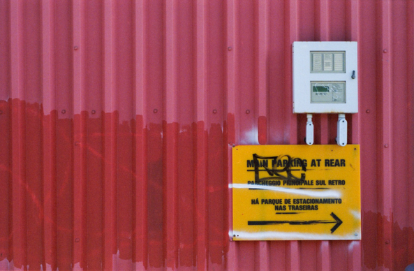 A faded red corrugated metal wall with an electrical box and a yellow sign that reads, under the graffiti, “main parking at rear”. The red has two tones: the original faded base and a fresher patch, presumably covering a graffiti tag.