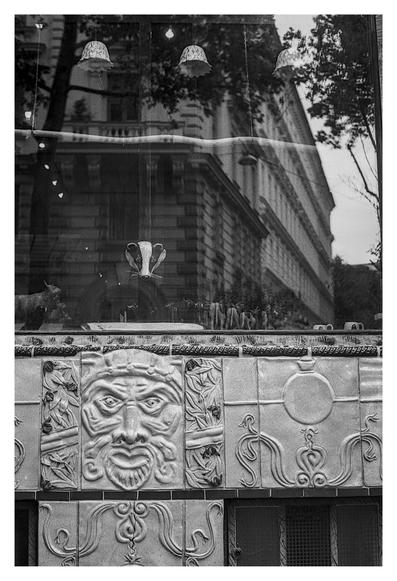Black and white photograph of a storefront window reflecting a grand historic building with ornate architecture and trees. Inside the window display, a sculpted badger head is visible among various objects. Below the window, decorative tiles feature a large, expressive face surrounded by intricate floral and serpentine designs.