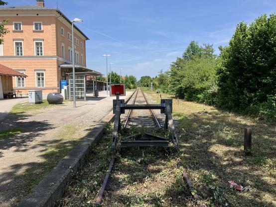 Blick am Prellbock vorbei auf ein einspuriges Bahngleis. Zur Linken ein Bahnhofsgebäude