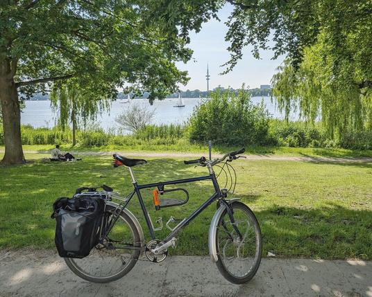 Ein Fahrrad steht auf dem radweg neben der Alster. Im Hintergrund die Alster mit Segelboot. Dahinter der Hamburger Fernsehturm.