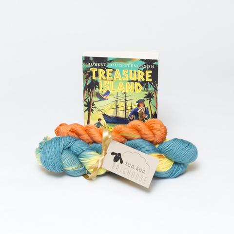 Picture shows a set of sock yarn consisting of a large skein which is blue and yellow with orange speckles and a mini skein which is orange. The two are tied together with gold ribbon. Behind the yarn stood on its end is a copy of the Puffin Classic edition of Treasure Island.