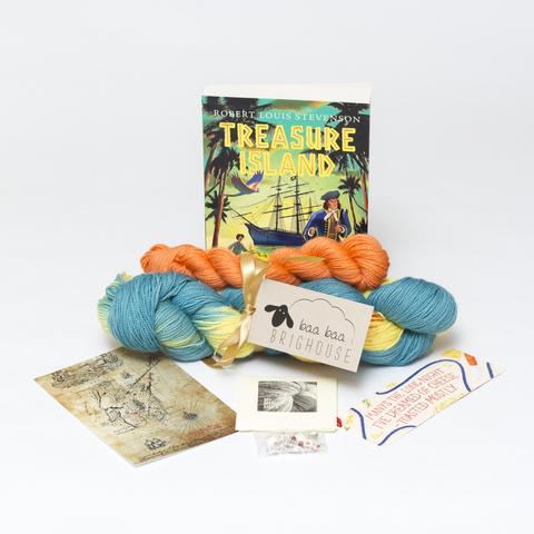 Picture shows a set of sock yarn consisting of a large skein which is blue and yellow with orange speckles and a mini skein which is orange. The two are tied together with gold ribbon. Behind the yarn stood on its end is a copy of the Puffin Classic edition of Treasure Island. In front of the yarn, from left to right, is a postcard, a packet of stitch markers and a bookmark.