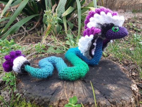 Dragon à plumes au crochet : corps en dégradé de bleu et vert, collerettes de plumes sur la tête, ailes et plumes sur la queue aux couleurs du drapeau ace : noir, gris, blanc et violet.
