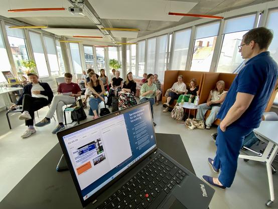 A group of about 20 people in a well-lit open office space sitting on chairs and sofas looking at a tall man standing next to a laptop computer that shows a slide on the Service Standard