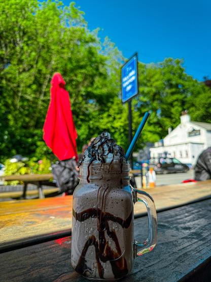 A chocolate milkshake in a jug with cream on top, and chocolate sauce. In the background, a road, a blue sign for the cafe, and some trees. The skies are blue and it’s a sunny day.