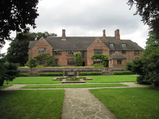 The image features a large, two-story brick house with a distinctive architectural style, surrounded by a well-maintained garden. The garden includes a central pathway leading to a rectangular area with plants or flowers and a landscaped lawn, contributing to an elegant outdoor setting. The sky is partly cloudy, and the surrounding trees add to the greenery and natural beauty of the scene.

Image Credits: Wikimedia / Bob Cantwell / CC BY-SA 2.0