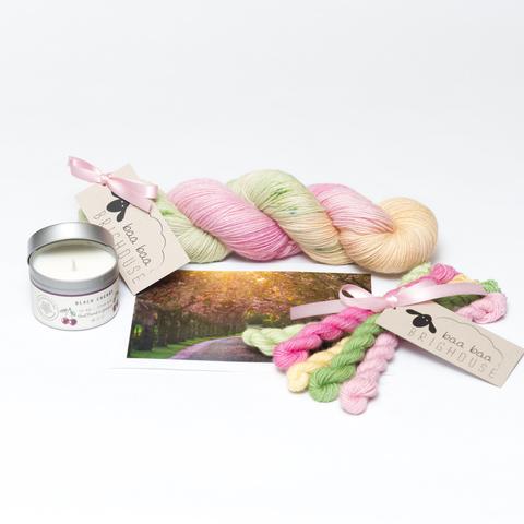 Picture shows a skein of yarn in shades of pink, green and yellow. In front of the yarn, from left to right, is a candle, a photography and a set of mini skeins.