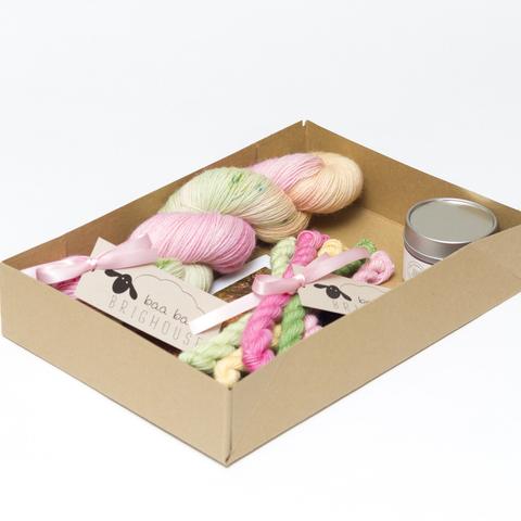 Picture shows an open A4 box containing a skein of yarn in shades of pink, green and yellow. A set of mini skeins in the smae colours can also be seen and a candle.