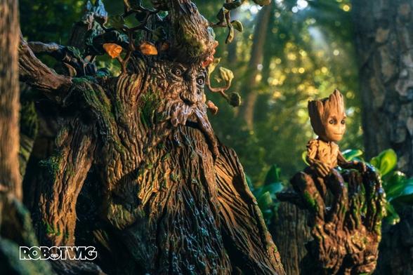 Treebeard holds Baby Groot in his hand whilst walking through Fangorn Forest. The sun beats on their faces and light can be seen coming through the foliage in the distance
