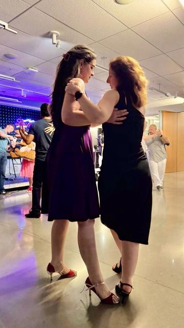 Two women in dresses and heels dance together. In the background, bandoneonist Heyni having a photo taken wirh a stuffed dinosaur.