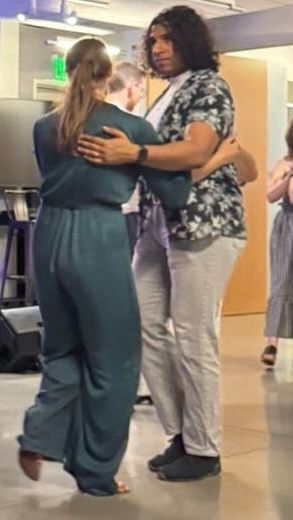 A person with a long blonde ponytail, wearing a blue jumpsuit, leads someone with shoulder-length curly hair wearing a floral shirt and khaki pants.