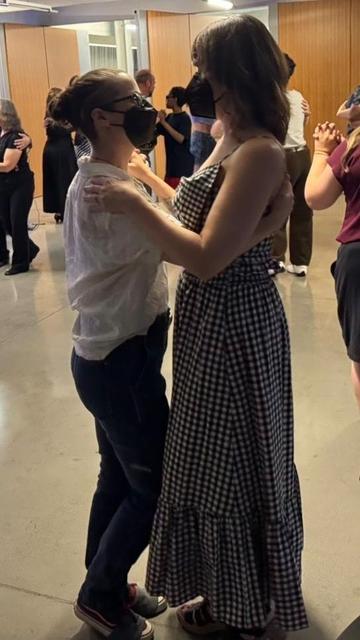 Two masked people dance together. One has a long checked dress and loose hair. The other has a white shirt, black pants, and hair in a bun.