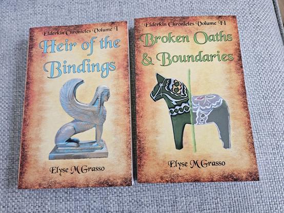 A photo of physical copies of the first two volumes of the Elderkin Chronicles. The sphinx faces right and the horse faces left, so they are looking at each other:

Heir of the Bindings: Elderkin Chronicles Volume I by Elyse M Grasso 
https://www.books2read.com/HeirOfTheBindings
Ebook ISBN: 978-1-966887-00-3
POD paper ISBN: 978-1-966887-01-0

A parchment look background with the series and author names in Black Chancery and the title in the same font but in blue. 

The main image on the cover is the winged Sphinx of Naxos as modeled by a souvenir figurine.

A fantasy set elsewhere in the multiverse. In a place not entirely unlike our Central Europe in 1700, an imperial Prince with eldritch ancestry faces threats and enemies, both material and otherwise, and begins to learn the truth about his heritage.

Broken Oaths & Boundaries: Elderkin Chronicles Volume II by Elyse M Grasso
https://books2read.com/BrokenOathsBoundaries
Ebook ISBN: 978-1-966887-02-7
Paper ISBN: 978-1-966887-03-4
A parchment look background with series and author names in Black Chancery and the title in the same font in green

Main image is a green Swedish Dala horse figurine with multicolored decorations, passing through a boundary marked by a green line. The tail end is less distinct and has distorted colors.

In an uncanny world a young Prince bound to Truth by eldritch ancestry must survive the schemes and oath-breaking of greedy Imperial Officials, and try to avert the hazards the oath-breaking evokes.