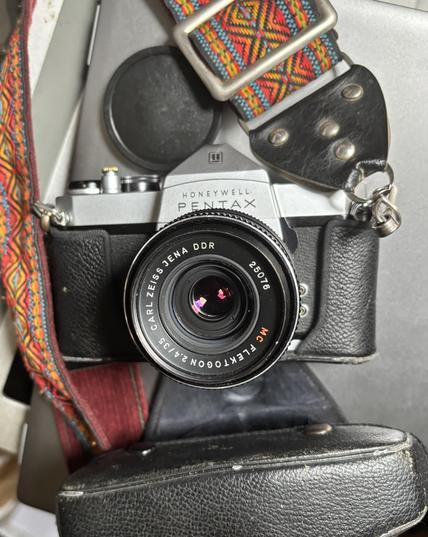 An old SLR camera, with a strap and leather camera case, but open. It’s a Pentax Spotmatic, but with a Zeiss lens (35mm focal length). The strap is orange and burgundy patterned. A lens cap is seen on the grey background.