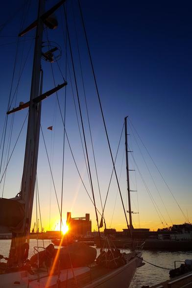 A striking vertical shot of a harbour at sunset, with the sun's warm glow reflecting on the water. In the foreground, the rigging and mast of a sailboat are silhouetted against the vibrant sky. Another sailboat's mast is visible to the right. In the background, the sun dips behind industrial buildings, casting a bright orange flare across the scene. The sky transitions from a deep blue at the top to a warm orange and yellow near the horizon.