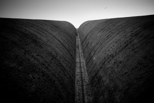 A black and white photograph shows a large cement structure with two massive, curved walls that form a V shape. The walls extend upward into the sky, with a narrow gap between them. In the sky above, a small bird can be seen