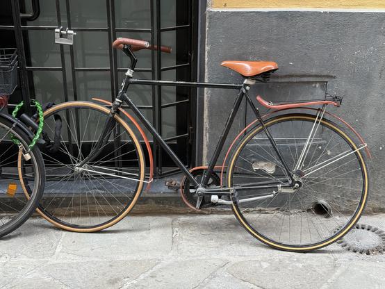A vintage black bicycle with a brown seat and handlebars is parked against a textured wall. The bike features thin tires with orange accents and is situated near a door and a grate on the ground.
