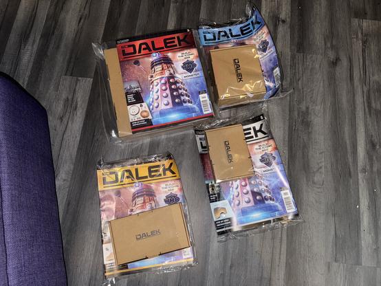 Several packages featuring "Dalek" from Doctor Who are arranged on a wooden floor. Each package includes a magazine and cardboard components for building a Dalek model. A purple cushion is visible in the corner.