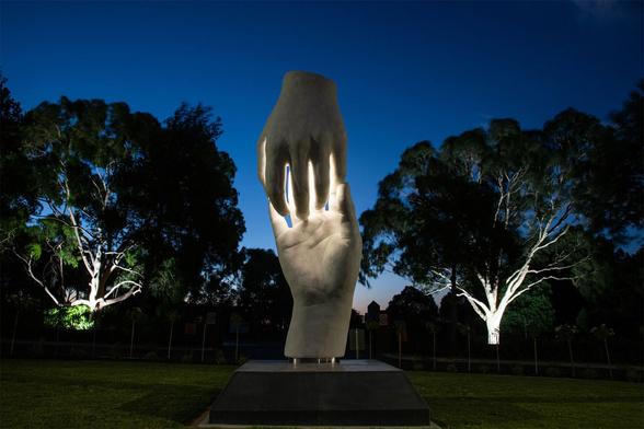 A large, illuminated sculpture of two hands, one reaching down to grasp the other, set against a twilight sky and surrounded by trees.