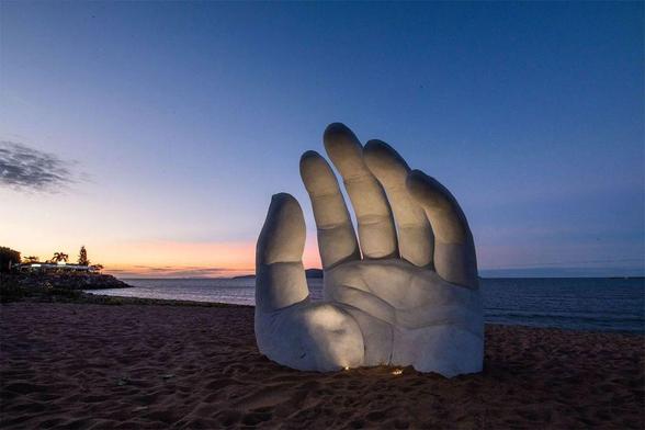 A large, white sculpture of a hand reaching up from the sandy beach, with a colorful sunset and calm ocean in the background.