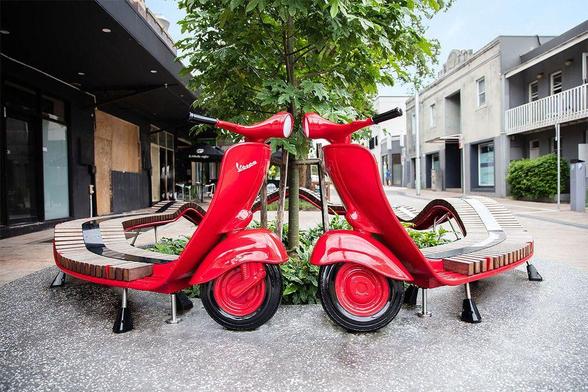 Two red Vespa scooter-inspired benches surround a small tree, set in a modern urban environment with nearby buildings.