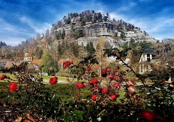 Das Bild zeigt eine idyllische Landschaft mit einem imposanten, steinigen Hügel im Hintergrund, der von Bäumen und Sträuchern bedeckt ist. Der Himmel ist blau mit einigen weißen Wolken. Im Vordergrund sind rote Rosen in voller Blüte zu sehen, die einen schönen Kontrast zur grünen Umgebung bilden. Im Hintergrund sind einige Gebäude mit roten Dächern zu erkennen, darunter ein Kirchturm, der auf eine kleine Stadt oder Gemeinde hinweist. Die Vegetation umfasst verschiedene Bäume und Sträucher, die in den Herbst übergehen, was durch die Farben der Blätter deutlich wird. Die Szene vermittelt ein Gefühl von Ruhe und natürlicher Schönheit.