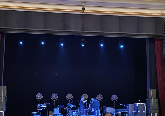 An empty, bluish lit stage. Oben an see some instruments and a few people building up things. There is a row of large lights in the back of the stage, towering over the instruments.