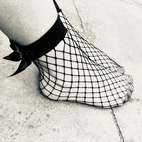 A photo of my feet from the right side. I’m wearing some fishnet socks that have a thick band at the ankle and black ribbon bows on the back of the band. My feet are resting on some paving and the photo is in greyscale.