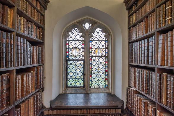 A medieval arched window with decorative stained glass is flanked by bookshelves filled with old, leather-bound books.