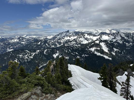Snowy mountain landscape with rugged peaks and dense evergreen forests. The sky is partly cloudy, and snow covers the slopes. Mt Rainier hidden in the clouds in the background.