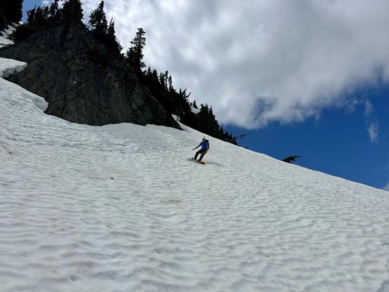 Snowboarding down a sun cupped snowy slope surrounded by rocky terrain and coniferous trees, under a partly cloudy blue sky.