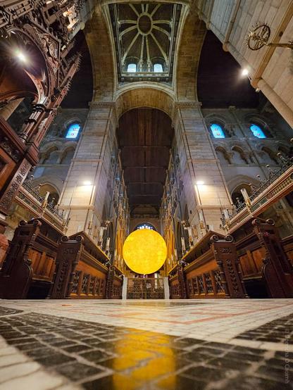 A wide-angle, low-angle photograph inside St Fin Barre's Cathedral, Cork, showing the ornate Gothic Revival architecture, wooden choir stalls, and stained glass windows. Dominating the centre is a large, glowing, spherical sculpture representing the sun, part of the Helios exhibition, suspended in the nave beneath the cathedral’s high vaulted ceiling.