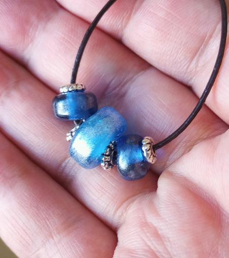 Image of a simple necklace of transparent blue glass lampwork beads enhanced with iridescent mica powder. Set against the artist's open hand.