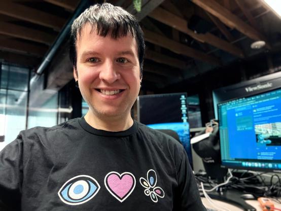 A photo of Gabe, wearing a tshirt with an eye, a heart, and the Owncast logo. He is surrounded by computer monitors.