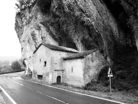 Fotografía en blanco y negro de una carretera en curva comarcal mojada, aunque no lleve en ese momento. Se ven unos árboles a lo lejos y el cielo está nublado. En primer plano hay pegada a la carretera una pequeña ermita que está construida contra la montaña que se eleva verticalmente sobre la carretera. En la ermita debe haber una santa, ya que hay un cartel fuera que pone “Virgen de Oskia”.
Antes hay una señal de peligro por desprendimiento de piedras en el borde de la carretera.