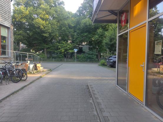 Eine schmale Durchfahrt zwischen zwei Gebäuden mündet in einer Einbahnstraße. Das Schild ist mittig von der Durchfahrt positioniert. Darunter hängt das Zusatzschild, dass Radfahren in beide Richtungen gestattet ist. Hinter dem Schild ist ein Zaun und ein Spielplatz.