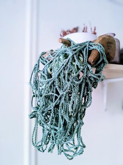 Photo d'une pièce d'art textile, un entrelacs fouillis de fils travaillés au crochet au point chaînette, dans les tons de bleu turquoise, suspendu à un morceau de bois posé sur une étagère