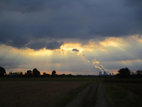 Biking along the fields back from Cologne, one afternoon a storm front with massive dark grey clouds cut the sky in half above me, the sunrays beaming out at the edges in white and golden yellow. 
A field path in the middle of the picture runs towards a steaming power plant at the horizon.