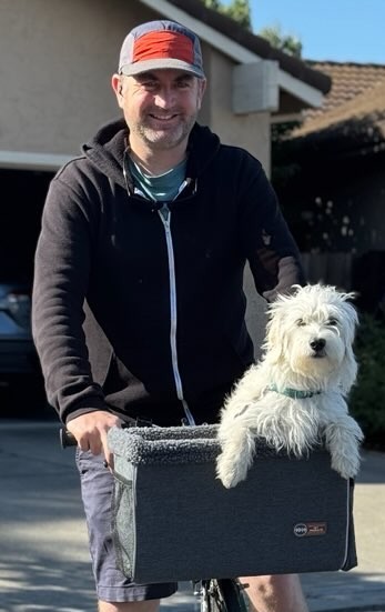 A white terrier sitting in a fleece-lined grey basket attached to a bike. A man wearing a black hoodie is standing stopped with the bike.
