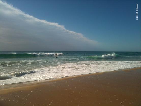A 2m swell on a green sea rolls in to a golden beach. Some surfers are catching the waves. Overhead, a pronounced cloud front with a distinct linear edge is advancing on the beach like a third wave. In front, the sky is blue.