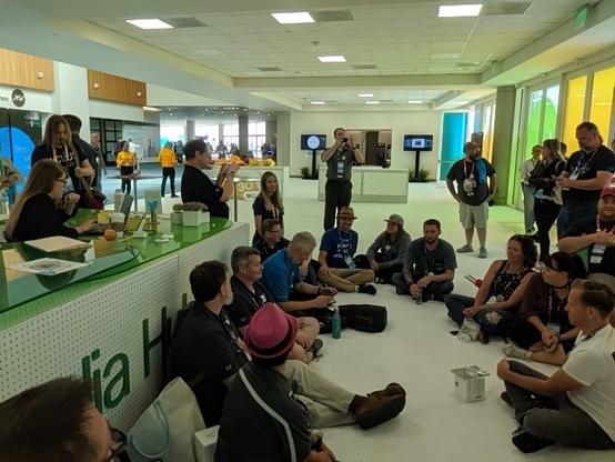 A group of people is seated on the floor in a spacious, brightly lit venue, engaging in a discussion. Some attendees are wearing event badges, and a woman at a table is speaking to the gathered audience. The background features a green and white counter