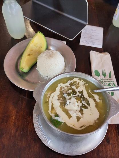Oven links eine kleine eckige Flasche Limonade. Auf einen flachen Blechteller ein Viertel Avocado und eine Portion Reis. In einer Blechschüssel grünliche Kartoffelsuppe mit einem Maiskolben, Kapern und Sahne oben auf. Rechts daneben liegt noch der Kassenzettel.