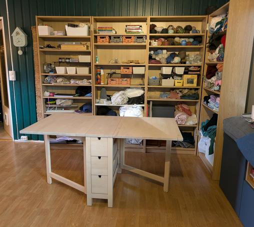 Bilde av et (noe rotete) sy-hjørne med et ikea slagbord foran 4 billy bokhyller med ymse håndarbeidsmateriale og verktøy.
