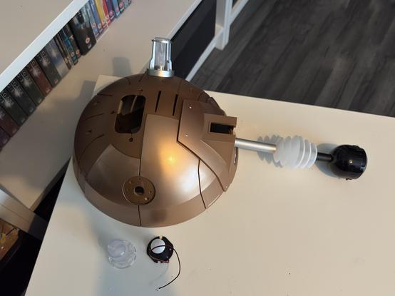 An incomplete model of a robotic character is shown, featuring a brown spherical body with various mechanical components, including a clear light atop the structure and a black attachment on a flexible arm. Additional small parts are placed nearby on a white table, with a background