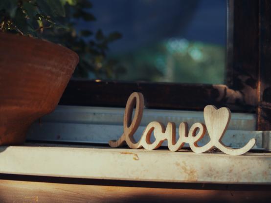 A decorative wooden cutout spelling the word “love” with a heart shape at the end rests on a sunlit windowsill. To the left, part of a terracotta plant pot is visible, and leafy green foliage can be seen as a reflection in the window.