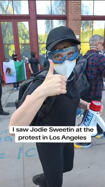 Jodie Sweetin out protesting in all black, eye pro, face mask, and a megaphone with a (LA)CAB sticker on it. She is giving a thumbs up.