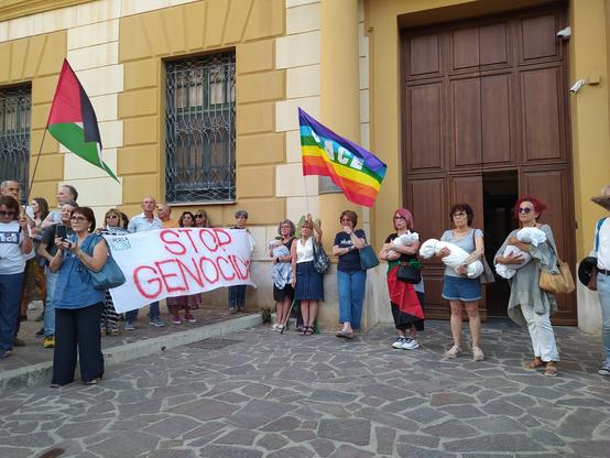 Persone di fronte all'ingresso della prefettura di Agrigento. Delle donne tengono in mano dei sudari per simboleggiare i bambini vittime delle bombe israeliane. Si vede uno striscione con la scritta 'stop genocide', una bandiera palestine e j a della pace