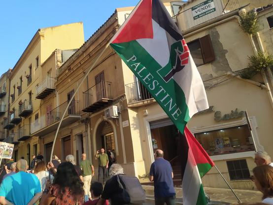 Persone di spalle. in primo piano una bandiera della Palestina con un pugno chiuso disegnato nel centro
