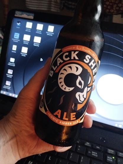 A bottle of Black Sheep Ale, held in my left hand with the label facing the camera. An open laptop with a Linux Mint xfce desktop behind.
