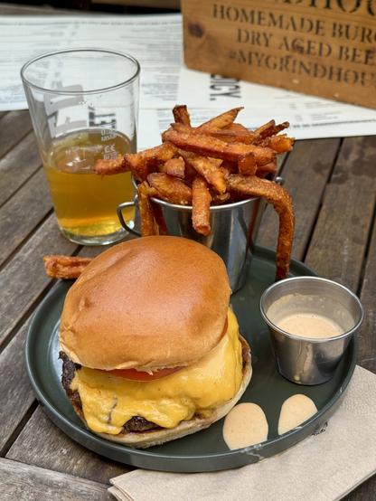 A cheeseburger with a melted cheddar slice and tomato sits on a plate, accompanied by a small cup of sauce. Next to it, a metal container holds crispy sweet potato fries, and a glass of beer is also present on the table.